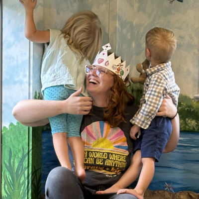 Children's librarian laughing wearing paper crown, holding 2 children