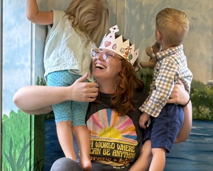 Children's librarian laughing wearing paper crown, holding 2 children