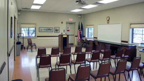 Community room with chairs, podium, ceiling-mounted projector, windows, and artwork