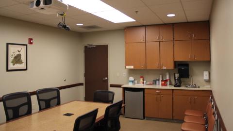 Conference room with table and chairs, ceiling-mounted projector, kitchenette, and artwork